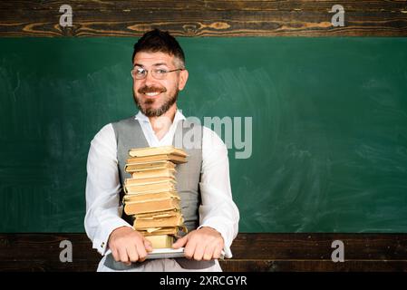 Lächelnder männlicher Student mit Notizbuch und Büchern vor der Tafel. Elektronische Bibliothek, E-Book. Bärtiger Lehrer mit Notizbuch und Lehrbüchern Stockfoto