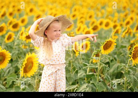 Süßes kleines Mädchen mit Sommerhut, das auf etwas im Sonnenblumenfeld zeigt Stockfoto