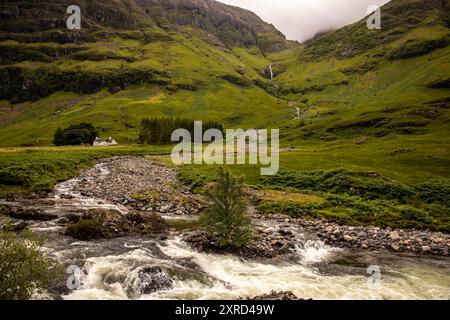 Glencoe, Schottland, Großbritannien. Juli 2024. Ferienhaus unter einem Wasserfall mit grünen Feldern und Schafweiden. Glencoe ist ein Dorf im Westen Schottlands. Er liegt im steilen Glencoe Valley in den schottischen Highlands. Die Gegend ist bekannt für Wasserfälle und Wanderwege, die Gipfel wie Buachaille Etive Mor und Bidean nam Bian erklimmen. Glencoe, ein ausgewiesenes National Scenic Area, das manchmal auch als „Glen of Weeping“ bezeichnet wird, war Schauplatz des berüchtigten „Massakers von Glencoe“ im Jahr 1692. (Kreditbild: © Ruaridh Stewart/ZUMA Press Press Wire) NUR REDAKTIONELLE VERWENDUNG! Nicht für kommerzielle ZWECKE! Stockfoto