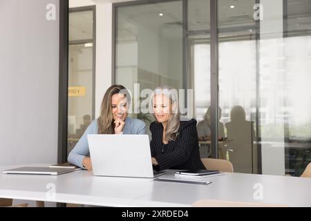 Positive jüngere und ältere Geschäftsfrauen, die Online-Inhalte auf einem Notebook ansehen Stockfoto