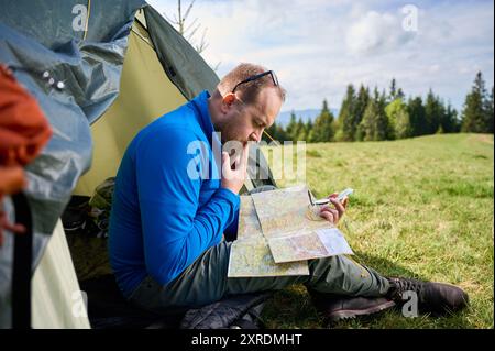 Der Mann sitzt im Eingang des Touristenzelt, tief auf die Karte fokussiert. Der Tourist hält den Kompass und denkt über seine Route nach, umgeben von üppigen grünen Feldern mit Bäumen und Bergen in der Ferne unter bewölktem Himmel. Stockfoto