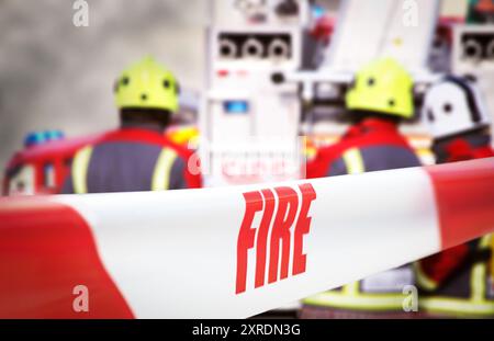 Mit rotem und weißem Band abgeriegelt, mit der Aufschrift „Feuer“. Im Hintergrund sind drei Feuerwehrleute und zwei Feuerwehrmaschinen unscharf Stockfoto