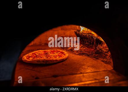 Pizza im Ofen mit brennendem Feuer backen. Pizza im Holzofen gekocht. Pizza, die Teig in einem Ofen mit offenem Feuer backt. Pizzakonzept. Vorbereiten Stockfoto