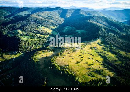 Blick aus der Vogelperspektive auf das sonnendurchflutete Tal mit kleinen Häusern und gewundenen Straßen, umgeben von dichten Wäldern. Goldenes Licht wirft lange Schatten. Die lebhafte Landschaft erstreckt sich über bewaldete Hügel. Stockfoto