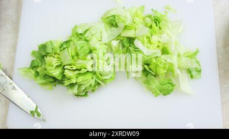 Frischer Eisbergsalat, der aus nächster Nähe auf weißem Küchenbrett geschnitten wird. Köcherhände in blauen Handschuhen hacken Grünkohl mit einem großen Eisenmesser. Kochen, Zubereiten von Zutaten in einem professionellen Restaurant. Ansicht von oben nach unten Stockfoto