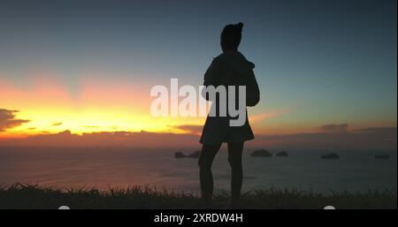 Eine Frau steht vor einem atemberaubenden Sonnenuntergang über dem Meer in einem thailändischen Resort. Der Himmel mit leuchtenden Orange-, Gelb- und Rosa-Tönen schafft eine dramatische Kulisse für diesen ruhigen Moment Stockfoto