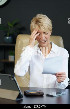 Eine Frau sitzt an ihrem Schreibtisch, sichtbar beunruhigt, während sie ein Dokument untersucht, umgeben von Bürobedarf und einer Topfpflanze. Stockfoto