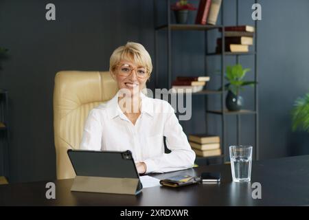 Eine lächelnde Frau in einem weißen Hemd greift an einem stilvollen Schreibtisch, umgeben von Büchern und einem erfrischenden Glas Wasser, mit ihrem Tablet in Berührung. Stockfoto