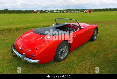 Klassischer Red Austin Healey 3000, der auf der Landebahn geparkt ist, mit Flugzeugen im Hintergrund Stockfoto