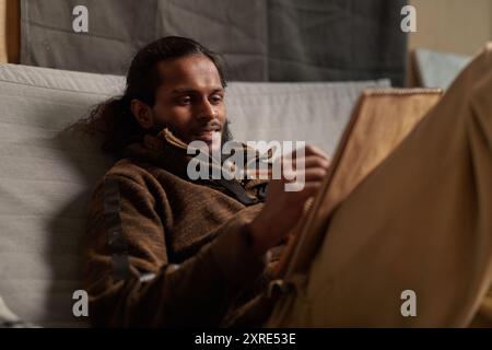 Bärtiger Mann, der auf einer grauen Couch sitzt und eine entspannte Haltung hat, während er auf einem Zeichenbrett skizziert. Er zeigt tiefen Fokus und künstlerische Konzentration Stockfoto