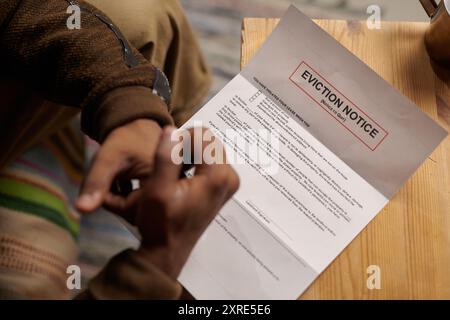 Person, die auf einem bequemen Stuhl sitzt und auf einem Holztisch einen Räumungsschein liest. Hände und Teilbetrachtung der Person sichtbar, wodurch ein Gefühl der Dringlichkeit entsteht Stockfoto