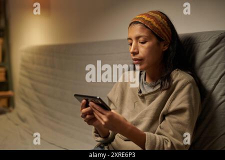 Hispanische Frau, die mit dem Smartphone auf dem Sofa sitzt und von einem schwach beleuchteten, gemütlichen Zimmer umgeben ist, legere Kleidung trägt und sich auf den Bildschirm konzentriert Stockfoto