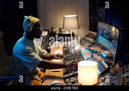 Mann, der an der Mischkonsole im Studio sitzt und den Audiopegel mit Konzentration anpasst. Umgeben von professioneller Audioausstattung und Computerbildschirmen, Kopfhörern und zielgerichtetem Ausdruck Stockfoto