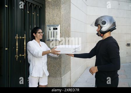 Ein Mann in einem schwarzen Hemd gibt einer Frau eine Pizza in einem weißen Hemd. Die Frau lächelt und der Mann trägt einen Helm Stockfoto