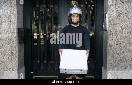 Ein Mann in einem Helm hält eine Pizzaschachtel in der Hand. Er lächelt und ist glücklich Stockfoto