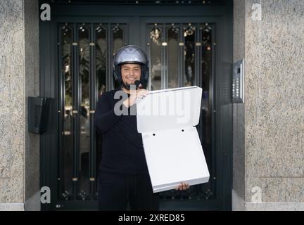 Ein Mann, der einen Helm trägt, hält eine Pizzaschachtel in der Hand. Er lächelt und ist glücklich Stockfoto