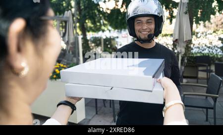 Ein glücklicher Mann, der einen Helm trägt, überreicht einer Frau eine Pizza. Stockfoto