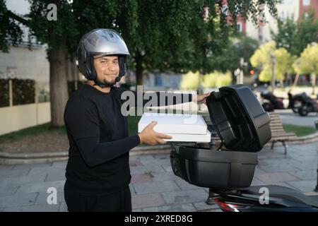 Ein Mann, der einen Helm trägt, hält eine Pizzaschachtel in der Hand. Er lächelt und ist glücklich Stockfoto
