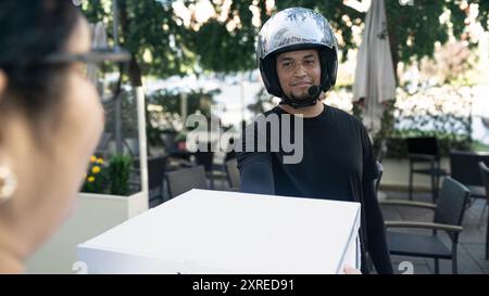 Ein Mann, der einen Helm trägt, gibt einer Frau eine weiße Box. Die Szene spielt in einem Restaurant mit mehreren Stühlen und Esstischen Stockfoto