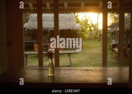 Tabanan, Bali, Indonesien - 9. August 2024: Blick auf einen grasbewachsenen Garten im Hinterhof und Sonnenuntergang vom Fenster einer hölzernen Hausterrasse mit einem rustikalen Tisch Stockfoto