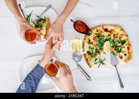 Ein Mann hält ein Mädchen an der Hand, während er frisch gekochte Pizza mit Getränken auf weißem Holztisch isst Stockfoto