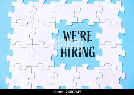 Ein Puzzle, in dem ein weißes Stück fehlt und die Worte Wir stellen ein, in Schwarz geschrieben. Das Puzzle ist auf blauem Hintergrund Stockfoto