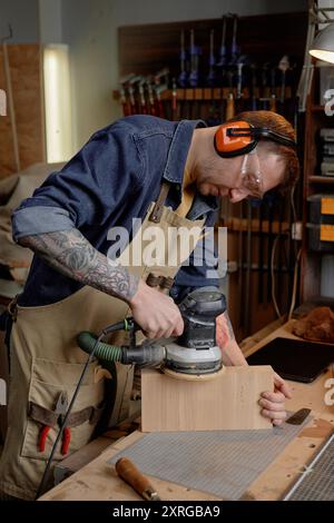 Mann, der in der Werkstatt elektrische Schleifmaschinen auf Holzblöcken verwendet, während er Schutzkleidung trägt und gesehen sich auf seine Aufgabe konzentriert. Hintergrund enthält Werkzeuge und Ausrüstung für Holzbearbeitungsprojekte Stockfoto
