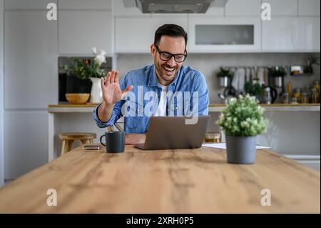 Fröhlicher junger Geschäftsmann, der die Hand winkt, während Videoanrufe über einen Laptop im Heimbüro Stockfoto