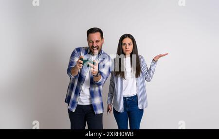 Wütende Freundin, die Gesten macht und in die Kamera schaut, während sie mit einem fröhlichen Freund steht und ein Videospiel spielt. Frustrierte Frau mit Happy Stockfoto