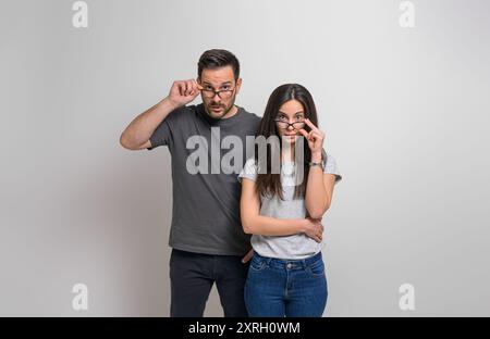 Schockierter junger Freund und Freundin, die durch eine Brille in die Kamera schauen. Enttäuschtes Paar, das in lässige gekleidet ist, starrt, während er isoliert steht Stockfoto