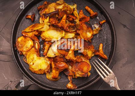 Gebratene Kartoffeln mit Pilzen Chanterellen in einer schwarzen gusseisernen Pfanne mit einer Gabel auf grauem Hintergrund. Hochwertige Fotos Stockfoto