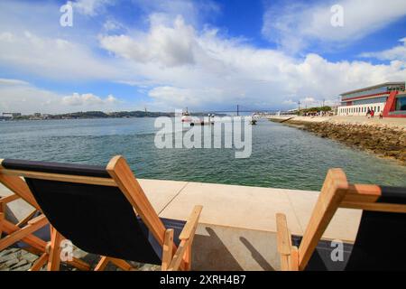 Lissabon, Portugal. Liegestühle am Fluss Tejo mit Blick auf die Brücke vom 25. April und die Christusstatue. März 2017 Stockfoto