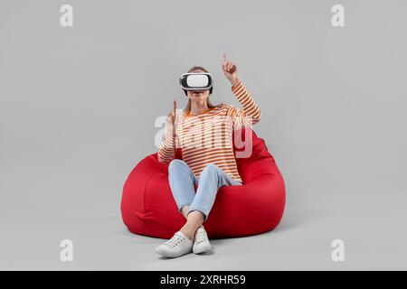 Glückliche Frau mit Virtual-Reality-Headset auf Sitzsack-Stuhl vor grauem Hintergrund Stockfoto