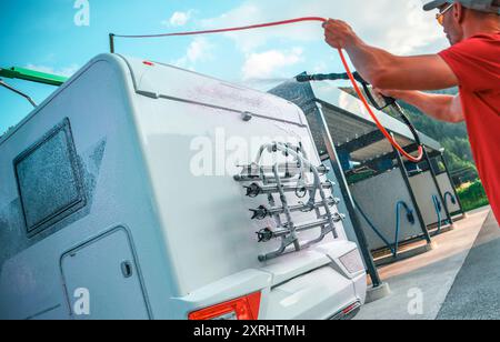 Ein Mann sprüht Wasser auf die Rückseite eines Wohnwagens, mit einem Schlauch an einer Waschanlage unter klarem blauem Himmel. Stockfoto