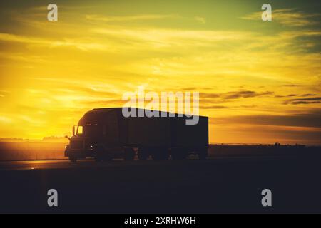 Bei Sonnenuntergang fährt ein Lkw eine offene Autobahn hinunter und wirft einen warmen goldenen Glanz über den Horizont und beleuchtet die Landschaft. Stockfoto