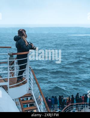 Antarktis - 26. Dezember 2023 - Foto des Eisbergs A23a vom Kreuzfahrtschiff hinter Menschenmenge. Passagier Mit Warmem Hut Hält Die Kamera Schaut Auf Riesiges Eis Stockfoto