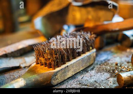 Nahaufnahme einer Metallwerkstatt mit Drahtbürste auf einer Werkbank, umgeben von Werkzeugen und Materialien in rustikaler Umgebung Stockfoto