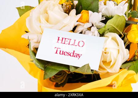 Die Worte "HELLO TUESDAY" auf einer weißen Visitenkarte vor einem Hintergrund frischer Blumen. Das Konzept von Liebe und Freude Stockfoto
