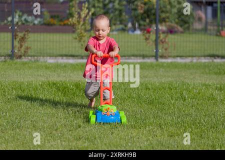 Das Kind mäht das Gras mit einem Spielzeug-Rasenmäher Stockfoto