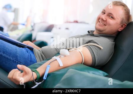Ein junger Mann sitzt bequem in einer medizinischen Einrichtung und spendet Blut während einer öffentlichen Veranstaltung. Er lächelt, während ein Arzt in der Nähe Geräte vorbereitet. Stockfoto