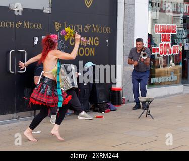 Mann spielt Geige, als Frau in Schottenrock tanzt auf der Prices Street während des Fringe Festivals in Edinburgh Schottland. August 2024 Stockfoto