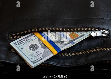 Band in Form der Flagge der Ukraine, gebunden auf einem Stapel von Hundert-Dollar-Scheinen in der Tasche eines schwarzen Mannes, finanzielle Hilfe für die Ukraine Stockfoto
