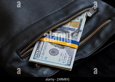 Band in Form der Flagge der Ukraine, gebunden auf einem Stapel von Hundert-Dollar-Scheinen in der Tasche eines schwarzen Mannes, finanzielle Hilfe für die Ukraine Stockfoto