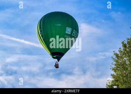 Grüner, sonnendurchfluteter Ballon in der goldenen Stunde, von der Bristol Balloon Fiesta 2024, als er über Bäume in der Nähe von Siston, Bristol, vorbeifährt. Am Samstag um 45 Uhr Stockfoto