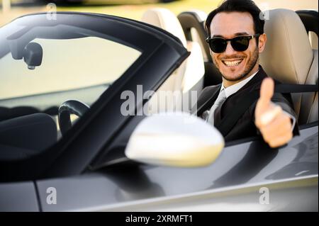 Ein fröhlicher junger Mann in Sonnenbrille und Anzug lässt die Daumen vom Fahrersitz eines Luxuskarosseries hochdrücken und zeigt Selbstvertrauen, Erfolg und ein Stockfoto