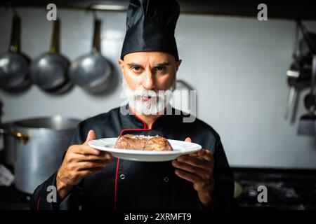 Professioneller Koch mit langem Bart in schwarzer Uniform, hält einen Teller mit einem köstlichen gegrillten Steak in einer Restaurantküche Stockfoto