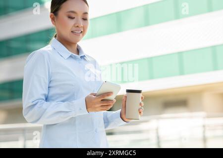 Frau, die Kaffee hält, während sie draußen auf dem Smartphone schreibt Stockfoto