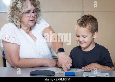 Ein kleiner Junge sitzt mit einem Lehrer an einem Tisch und schreibt mit Markierungen. Der Lehrer beobachtet den Jungen und scheint ihm bei seiner Arbeit zu helfen. Die Stockfoto