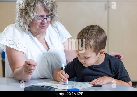 Ein kleiner Junge sitzt mit einem Lehrer an einem Tisch und schreibt mit Markierungen. Der Lehrer beobachtet den Jungen und scheint ihm bei seiner Arbeit zu helfen. Die Stockfoto