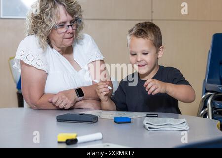 Ein kleiner Junge sitzt mit einem Lehrer an einem Tisch und schreibt mit Markierungen. Der Lehrer beobachtet den Jungen und scheint ihm bei seiner Arbeit zu helfen. Die Stockfoto